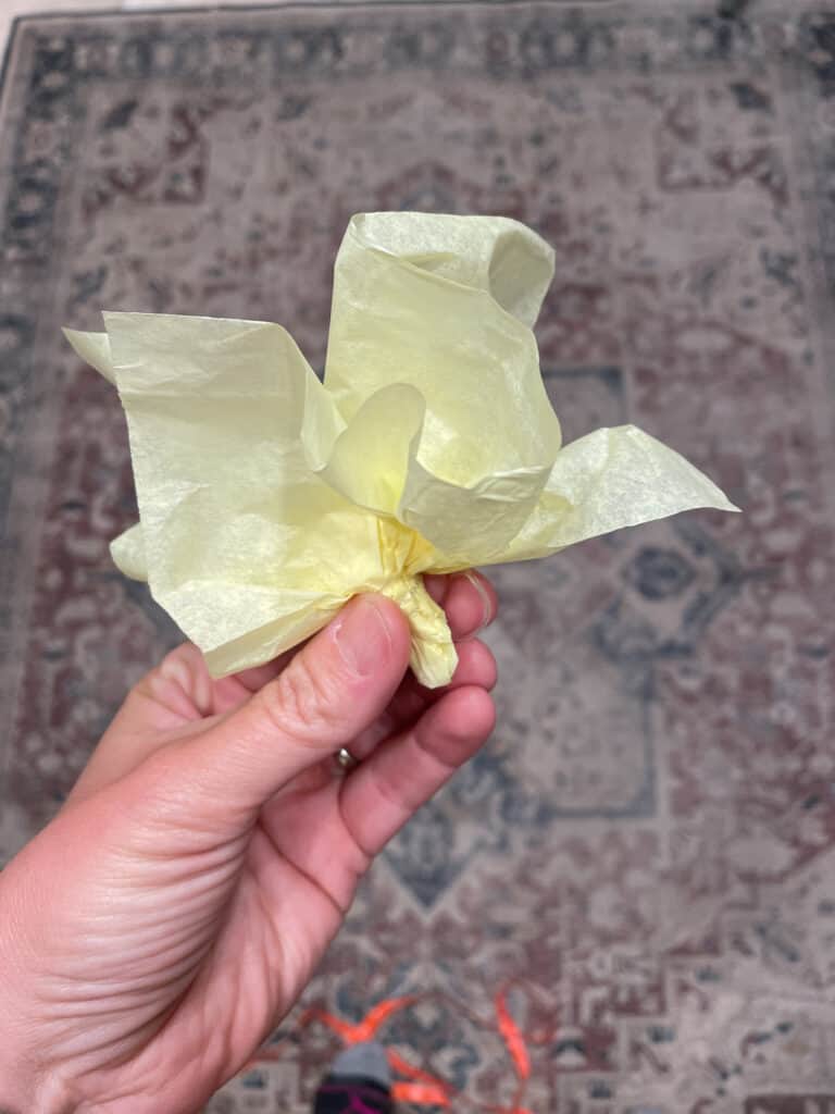 yellow tissue paper being held  to demonstrate how to bunch the tissue paper up and secure it with clear elastic band to create the popcorn kernel