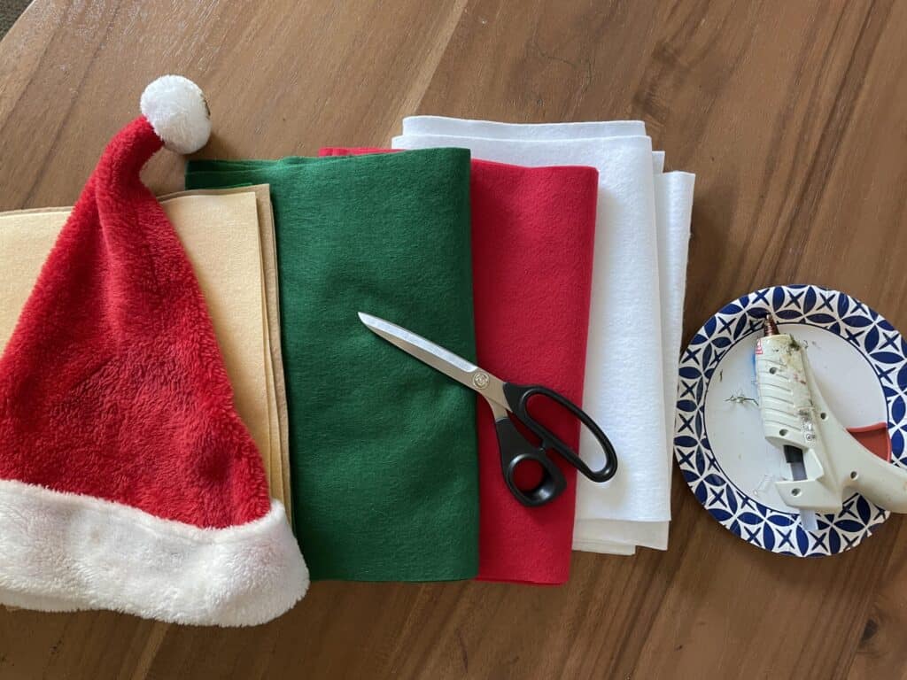 Red and white santa hat laid on top of nude, green, red and white pieces of felt.  Scissors and a hot glue gun are on the side.