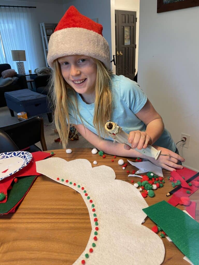 Child wearing santa hat holding hot glue gun and embellishing a white felt elf collar with colorful pom poms.